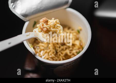 Une tasse de nouilles instantanées avec une fourchette en plastique. Les nouilles sont dans un bouillon avec des morceaux de viande et des légumes. Banque D'Images