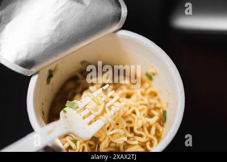 Une tasse de nouilles instantanées avec une fourchette en plastique. Les nouilles sont dans un bouillon avec des morceaux de viande et des légumes. Banque D'Images