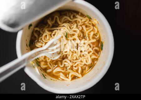 Une tasse de nouilles instantanées avec une fourchette en plastique. Les nouilles sont dans un bouillon avec des morceaux de viande et des légumes. Banque D'Images