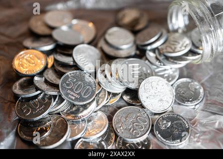 Un tas de pièces de monnaie indonésiennes de 500 roupies dispersées sur une surface métallique avec quelques pièces de monnaie débordant d'un bocal en verre. Banque D'Images