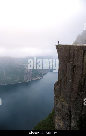 Un seul individu se dresse au bord de Preikestolen, une falaise en Norvège surplombant un fjord. Le ciel est brumeux et le paysage est dramatique. Preikestolen, Norvège Banque D'Images