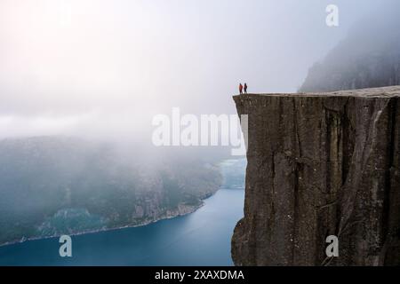 Une vue depuis le sommet du Pulpit Rock en Norvège, avec deux personnages debout sur le bord, regardant au-dessus du fjord brumeux. Preikestolen, Norvège Banque D'Images