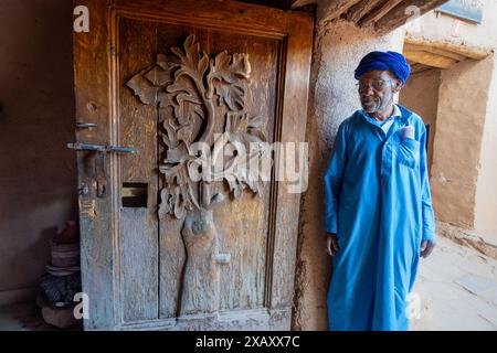 Ait Ben Haddou, Maroc - 19 mars 2024 : visite de maisons berbères traditionnelles. Debout à côté d'une porte en bois magnifiquement sculptée était un vieil homme portant Banque D'Images