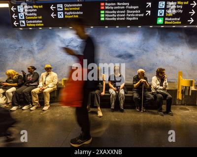 Les voyageurs attendent le prochain train à la station de métro T-Centralen, le centre névralgique du réseau de métro de Stockholm. L'artiste Per Olof Ultvedt a conçu cette zone pour l'ouverture de la ligne bleue en 1975, dans son bleu signature avec des vignes florales stylisées peintes sur le plafond de roche brute. Un passager portant un sac orange dépasse rapidement le banc. Le panneau suspendu indique les sorties, les liaisons de bus et d'aéroport, les trains de banlieue et plusieurs lignes de métro. Avec environ 100 stations conçues de manière artistique, le métro de Stockholm est considéré comme la plus longue galerie d'art du monde. Klara, Stockholm, Comté de Stockholm, Suède. Banque D'Images