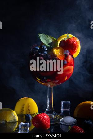 Cocktail fruité dans un verre à vin, avec des fraises fraîches, des bleuets et des oranges, sur un fond sombre avec de la fumée Banque D'Images