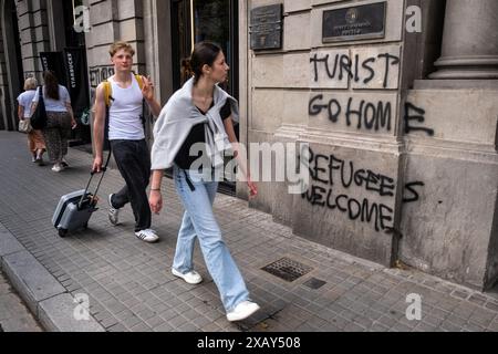 Barcelone, Espagne. 09 juin 2024. Un jeune couple de touristes est vu passer devant le graffiti anti-tourisme. Comme la plupart des capitales européennes, Barcelone vit entre la nécessité d'une économie touristique dont elle ne peut se passer et le rejet d'un grand nombre de citoyens contre la distorsion culturelle et la précarité de l'emploi typiques de l'industrie touristique. Les slogans sur les espaces publics de la dernière manifestation contre la surpopulation touristique coexistent avec une activité touristique qui ne se repose jamais. (Photo Paco Freire/SOPA images/SIPA USA) crédit : SIPA USA/Alamy Live News Banque D'Images