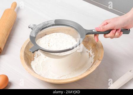 Femme tamisant la farine dans le bol à la table en bois blanc dans la cuisine, gros plan Banque D'Images