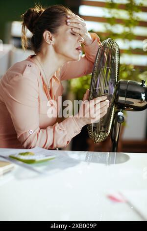 Lieu de travail durable. ouvrière moderne dans un bureau vert moderne avec ventilateur électrique souffrant de la chaleur estivale. Banque D'Images