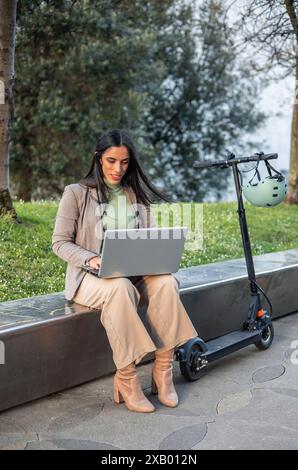 femme d'affaires verticale est assise sur un banc à côté de son scooter électrique, concentré sur son ordinateur portable. scene met en valeur le mélange moderne de mobilité et tec Banque D'Images