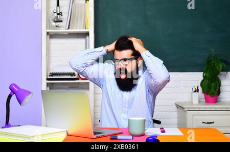 Étudiant universitaire masculin en colère travaillant avec un ordinateur portable en classe. ELearning et éducation. Professeur barbu dans des lunettes criant assis à la table wi Banque D'Images