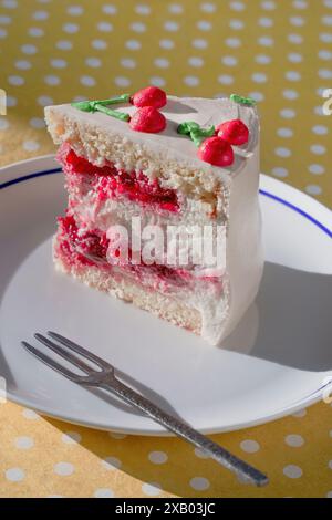 Gros plan d'une tranche de gâteau de couche de cerise avec glaçage blanc et décorations de cerise sur une assiette blanche. Nappe à pois et fourchette à dessert dans le bac Banque D'Images