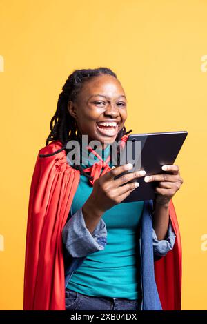 Fille ravie de rire tout en portant le costume de super-héros pour Halloween, tenant la tablette, s'amusant, fond de studio. Femme extatique excitée d'être habillée en héros de bande dessinée, à l'aide de l'appareil Banque D'Images