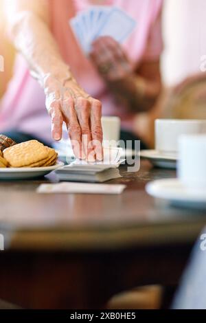 Femme âgée, table et main avec des cartes dans la maison de retraite pour l'activité, le passe-temps et le plaisir le week-end. Jeux, personne senior et se détendre avec snack in Banque D'Images