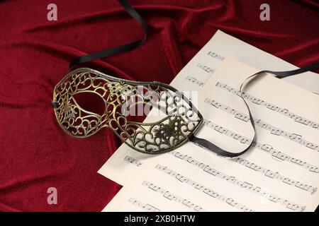 Masque facial élégant et feuilles de musique sur tissu rouge. Représentation théâtrale Banque D'Images