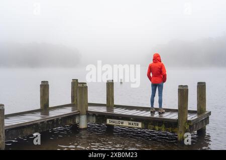 Dame en manteau de pluie rouge de derrière surplombant la vue brumeuse après la randonnée autour d'un lac Banque D'Images