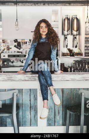 Belle barista féminine travaillant dans un café, assise sur le comptoir. Étudiant universitaire travaillant à temps partiel dans un café. Banque D'Images