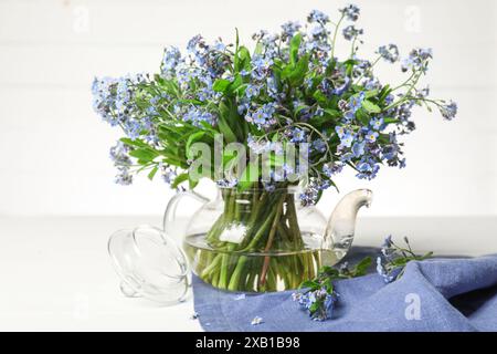 Bouquet de belles fleurs oubliées dans une théière en verre et un tissu bleu sur une table blanche Banque D'Images