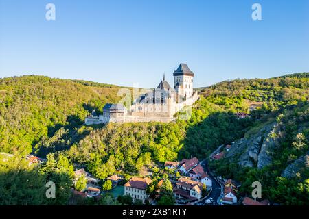 Le château de Karlstejn se dresse au sommet d'une colline, entouré de forêts vertes denses sous un ciel bleu clair, avec un village pittoresque niché dans la vallée en contrebas. République tchèque Banque D'Images