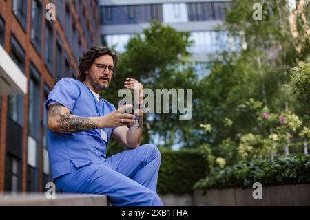 Travailleur de la santé prenant une pause, fumant une cigarette à l'extérieur et défilant au téléphone, devant le bâtiment de l'hôpital. Banque D'Images