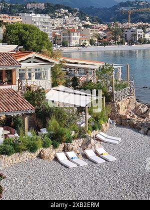 Une ville côtière pittoresque avec un coin salon en bord de mer serein avec des chaises longues blanches sur une plage rocheuse. Les charmantes maisons et la verdure luxuriante blen Banque D'Images