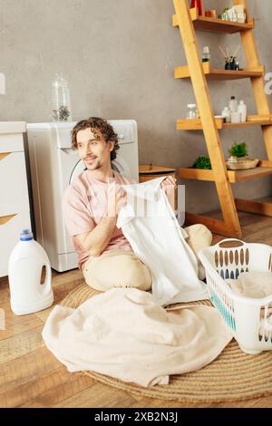 Un homme dans des vêtements de maison confortables assis sur le sol, pliant chemise blanche. Banque D'Images