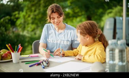 Talentueuse petite fille dans un Jumper jaune dessinant de belles images avec une bénévole d'une organisation caritative locale. Travailleur de l'aide humanitaire aidant l'enfant avec les devoirs de cours d'arts. Banque D'Images