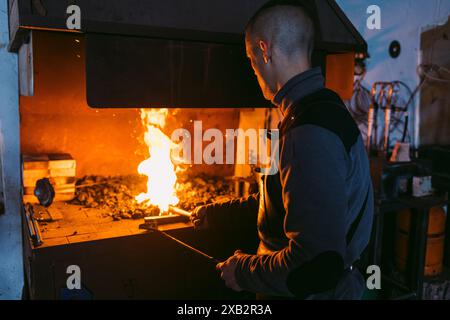 Vue arrière de forgeron masculin méconnaissable travaille avec une concentration intense, chauffant la ferronnerie dans les braises incandescentes d'une forge traditionnelle. Banque D'Images