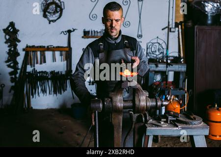 Un forgeron masculin concentré travaille à façonner du métal chaud avec un marteau sur l'enclume dans son atelier, entouré d'outils traditionnels de forgeron. Banque D'Images