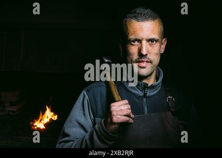 Forgeron masculin confiant avec marteau dans une forge debout regardant la caméra. Banque D'Images