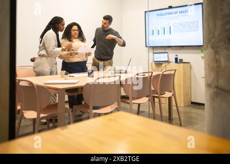 Une équipe diversifiée de professionnels collabore sur des documents et des projections de données sur un écran dans un espace de coworking lumineux Banque D'Images