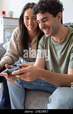 Un jeune couple joyeux s'assoit à proximité sur un canapé, partageant un moment léger tout en jouant à des jeux vidéo. Leurs expressions montrent amusement et affection Banque D'Images