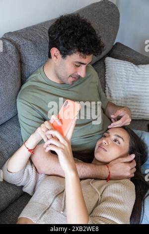Un jeune couple profite d'un moment confortable sur un canapé, avec la femme tenant un smartphone, à la fois apparemment engagée et heureuse Banque D'Images