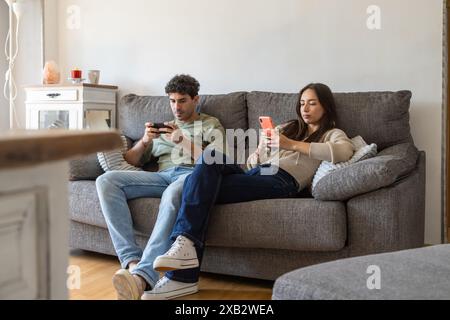 Un jeune homme et une jeune femme assis côte à côte sur un canapé gris, chacun immergé dans son propre smartphone, représentant la vie domestique moderne et le rôle de Banque D'Images