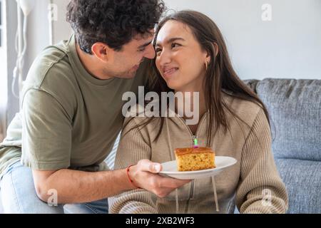 Un jeune couple joyeux partage un rire et un morceau de gâteau dans leur salon confortable, reflétant une scène chaleureuse et domestique Banque D'Images