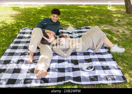 Un jeune couple profite d'un moment de sérénité ensemble, allongé sur une couverture de pique-nique noir et blanc à carreaux dans un parc luxuriant, avec des accessoires de style de vie Banque D'Images