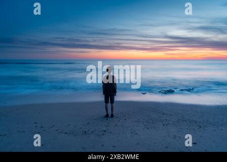 Un individu isolé méconnaissable se dresse sur les rives sablonneuses de Atlanterra Beach, absorbé par le coucher de soleil à couper le souffle sur l'horizon. Banque D'Images