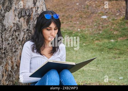 Une image sereine capturant une femme prise dans un livre tout en s'appuyant contre un arbre dans un cadre paisible de parc Banque D'Images