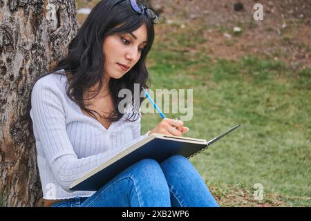 Une femme pensive est assise sous un arbre, immergée dans son cahier, entourée par la tranquillité d'un parc luxuriant, exprimant créativité et détente Banque D'Images