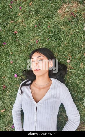 Une jeune femme est allongée sur l'herbe les yeux fermés, profitant d'un moment paisible entouré par la nature dans un cadre paisible de parc Banque D'Images