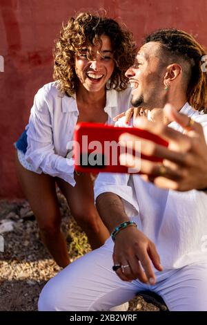 Un jeune couple joyeux dans une tenue blanche décontractée profite d'un moment ludique tout en prenant un selfie contre un mur rouge vif à l'extérieur la femme rit vif Banque D'Images