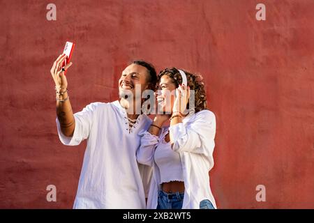 Un jeune couple joyeux aime prendre un selfie avec un smartphone rouge, souriant largement sur un fond rouge vif les deux portent des tenues décontractées blanches et Banque D'Images