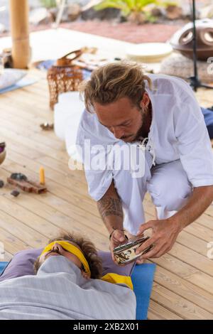 Un homme effectue une thérapie sonore sur un autre individu en utilisant des bols chantants tibétains pendant une séance de bain de gong en plein air. Les deux participants ne sont pas reconnaissables Banque D'Images