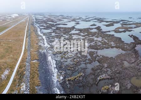 Vue aérienne d'un paysage islandais tranquille avec une route sinueuse le long d'une côte brumeuse, parsemée de piscines naturelles et d'un terrain mousselé. Banque D'Images