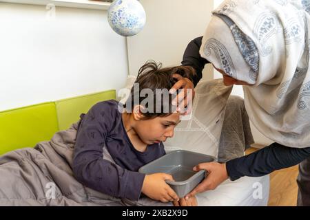 Un jeune garçon souffrant d'hypoglycémie est réconforté par sa mère anonyme, qui porte un hijab, alors que l'enfant tient un bol malade. Banque D'Images