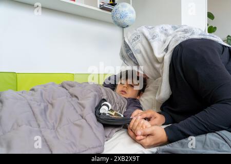 Vue de côté d'une mère musulmane méconnaissable avec le hijab tient la main de son jeune fils alors qu'il est allongé dans le lit et malade, avec un appareil médical à proximité, suggérant Banque D'Images