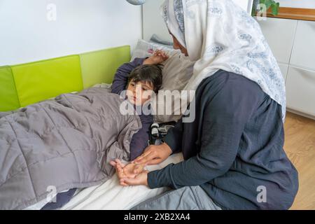 Vue de côté d'une mère musulmane méconnaissable avec le hijab tient la main de son jeune fils alors qu'il est allongé dans le lit et malade, avec un appareil médical à proximité, suggérant Banque D'Images