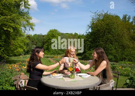 Trois amis partagent le rire et le café à une table ronde dans un jardin verdoyant, entouré d'arbres et de fleurs Banque D'Images