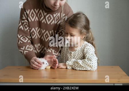 Une mère recadrée méconnaissable et sa fille manipulent doucement un hamster blanc moelleux à une table en bois, concentrés sur l'animal avec les yeux ouverts, shar Banque D'Images