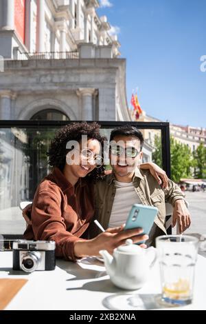 Un joyeux couple multiethnique, un Chinois et une femme hispanique, prenant un selfie dans un café de Madrid, en Espagne. Le paysage urbain emblématique et une caméra sont v Banque D'Images