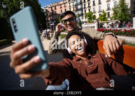 Un couple multiethnique, composé d’un Chinois et d’une femme hispanique, profite d’une journée ensoleillée à Madrid, en Espagne. Ils sont assis sur un banc portant des têtes Banque D'Images
